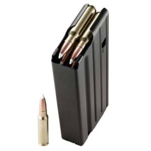 Black plastic ammunition magazine partially filled with rifle cartridges, with a single cartridge lying on the white surface beside it.