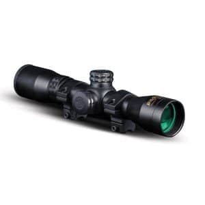 Black firearms scope with green-tinted objective lens and mounting rings on a white background, photographed from the side.
