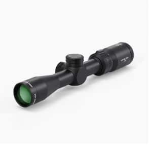 Black telescopic rifle scope with green-tinted front lens and adjustable dials along the tube, lying on a white surface to the right of center.