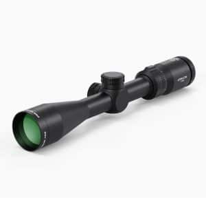 Black riflescope with a green-tinted objective lens and exposed adjustment turrets on a white background to the right.