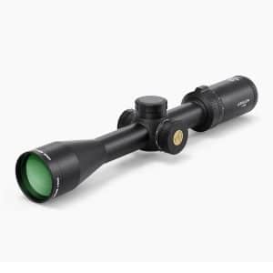 Black riflescope with green-tinted objective lens, centered adjustment turrets, and knurled eyepiece shown at an angle on a white background